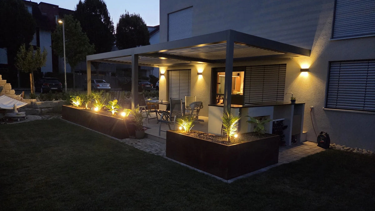 Freistehendes BUDDY Lamellendach SB 400 auf einer Kundenterrasse am Abend, Lamellen geschlossen, beleuchtete Pflanzbeete